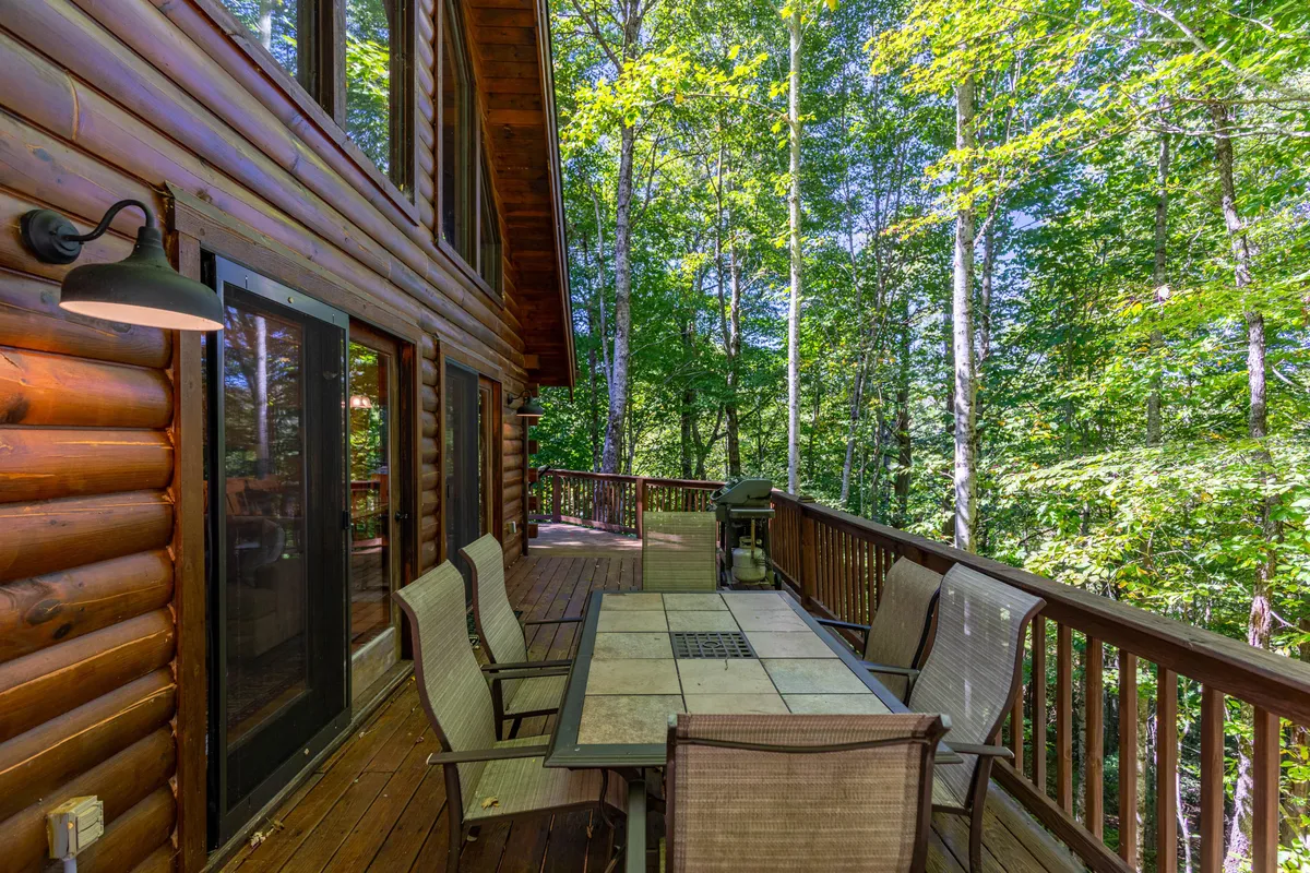A spacious wooden deck features a large outdoor dining table, surrounded by trees and complemented by a BBQ grill. The setup is ideal for group gatherings, outdoor dining, and sunset dinners beneath the sky, illustrating the deck's versatility and the BBQ amenity.