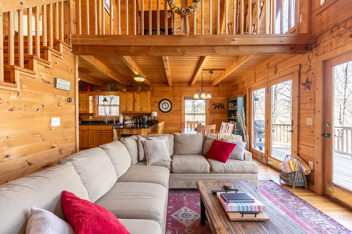 A spacious open-plan living and dining area, featuring a large sectional sofa, a wooden coffee table, expansive windows flooding the room with natural light, and a visible dining table that seats six. The inviting layout encourages gathering, relaxation, and shared activities, with a cozy, rustic ambiance enhanced by warm wood accents.