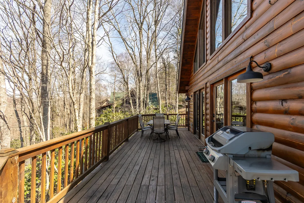 A cozy wooden deck set among trees, featuring a grill, outdoor seating, and open space for gatherings. The inviting atmosphere is perfect for barbecues, morning coffee, or evening relaxation with friends and family.