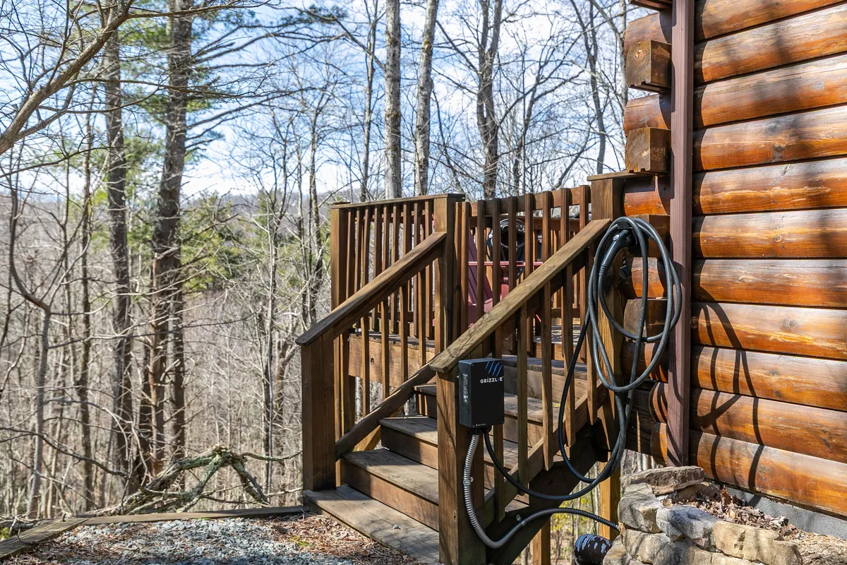 A wooden deck with steps leading up to the log cabin exterior, nestled among tall trees. The scene evokes an inviting transition between the cottage and nature, perfect for morning coffee or post-adventure gatherings.