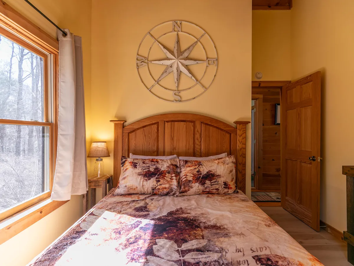A well-lit bedroom with patterned bedding, a wooden headboard, compass wall décor, and a window streaming in natural light. The setting is airy and designed for relaxation.