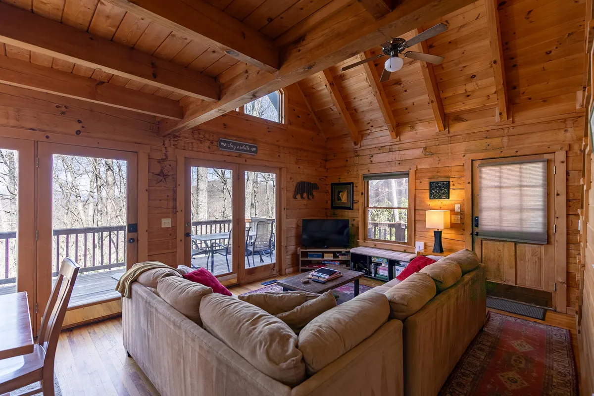 A spacious living area featuring a large couch, rustic wooden beams, windows overlooking natural scenery, and a TV. The space is well-lit, comfortable, and ideal for both relaxation and group activities.