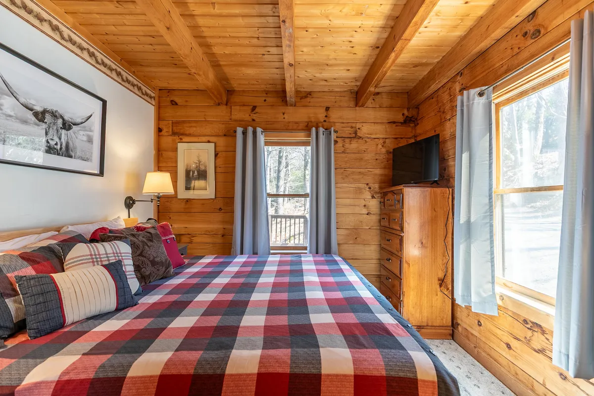 A welcoming bedroom with a plaid bedspread, prominent wooden beams, and a large window looking out onto nature. The setup evokes warmth and comfort, ideal for relaxation and privacy.
