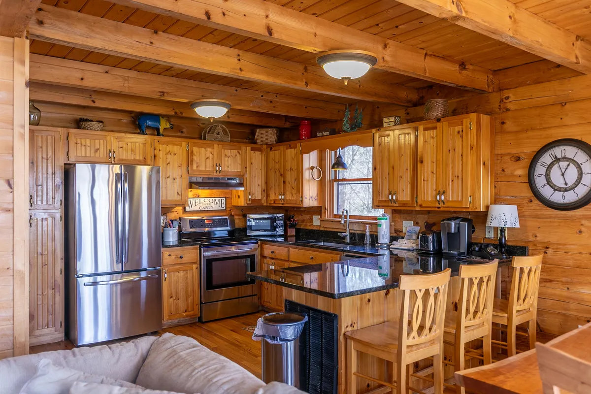A bright, welcoming kitchen with stainless steel appliances, black granite countertops, warm wooden cabinetry, and a large window overlooking the outdoors. The space is set up for cooking and gatherings, with modern amenities seamlessly integrated into the rustic charm of the cottage.