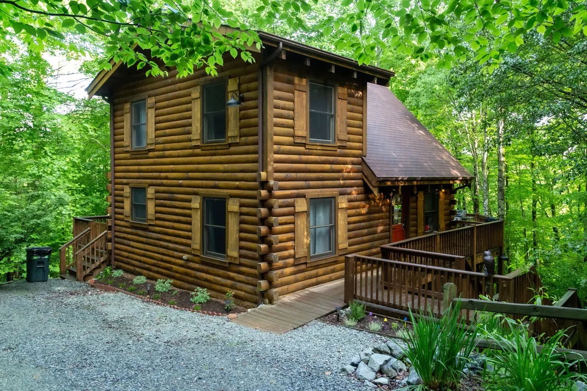 A picturesque log cabin surrounded by lush greenery, featuring a spacious deck perfect for relaxing, with forest views that set a tranquil, cozy mood. The cabin's rustic charm is enhanced by its inviting porch, ideal for enjoying sunsets, listening to the soothing sounds of nature, and stargazing at night.