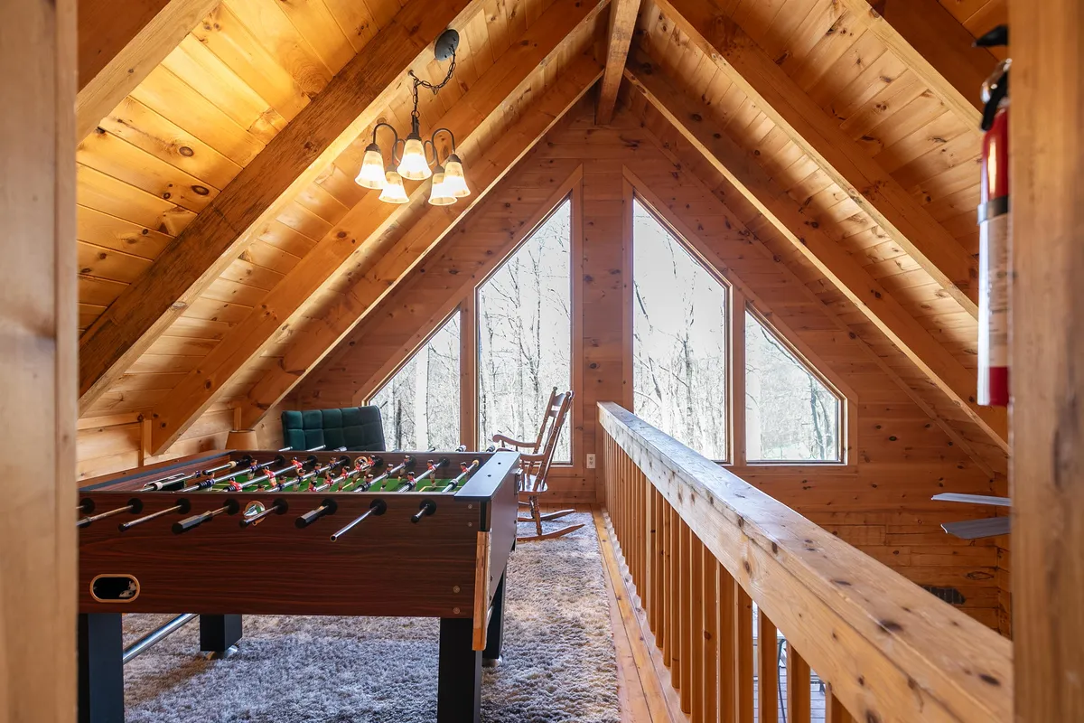 A bright, playful loft space with a foosball table set in front of large windows, surrounded by wooden railings and a cozy rocking chair that overlooks nature. The area is bathed in natural light, creating an inviting spot for games or relaxation.