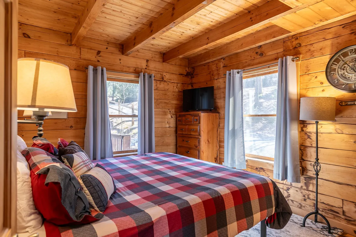 This image features a cozy bedroom with a plaid bedspread, warm wooden beams, and large windows allowing natural light to filter in. A table lamp and dresser add to the homey, restful atmosphere.