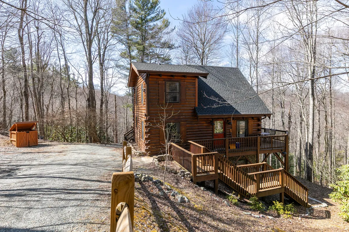 This image captures the log cabin structure of Mimi's Cottage set amidst tall trees, with a spacious wooden deck in the foreground. The deck features comfortable seating, and the natural surroundings suggest a peaceful retreat just minutes from local attractions.