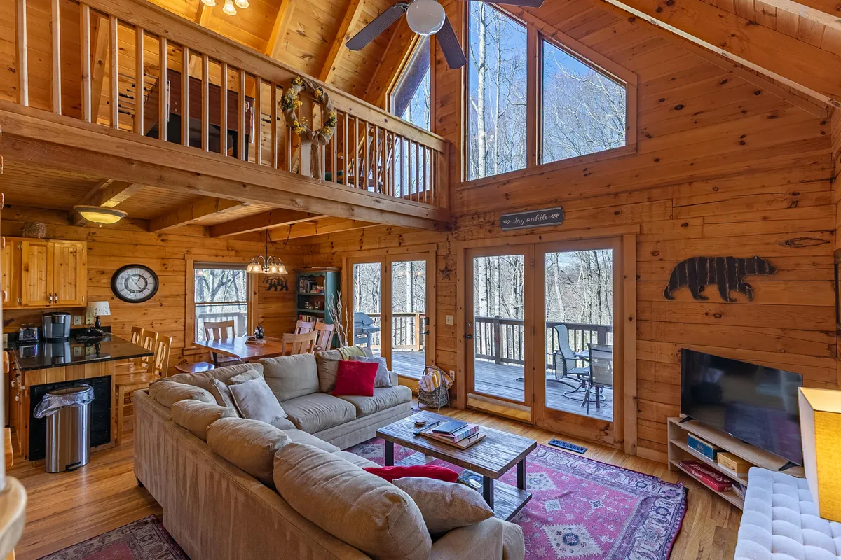 A cozy, sun-filled living and dining area with large windows, a comfortable sofa, a wooden dining table, and a decorative bear silhouette on the wall. The space feels open and welcoming, with natural light pouring in.