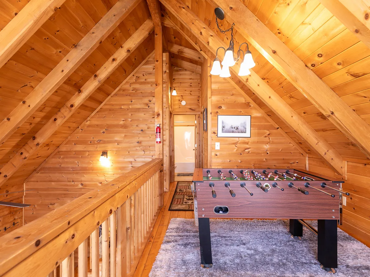 A casual, fun-filled interior view centered on a foosball table beneath rustic wooden beams and a chandelier. The open space is ideal for friendly tournaments and socializing, reinforcing the playful side of the entertainment retreat.
